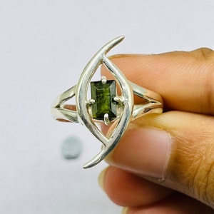 May include: A silver ring featuring a green gemstone set in a distinctive, abstract design. The ring has two curved lines that meet to form a shape reminiscent of an eye.
