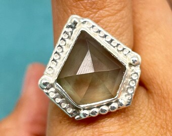 May include: A silver ring with a faceted gray gemstone set in a geometric design. The ring has a beaded border.