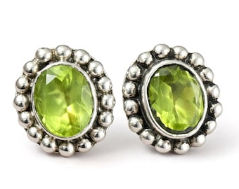 May include: A pair of stud earrings featuring oval-cut, light green peridot gemstones set in sterling silver. Each earring is surrounded by a decorative border of small, polished silver spheres, creating a textured, beaded frame around the stone.