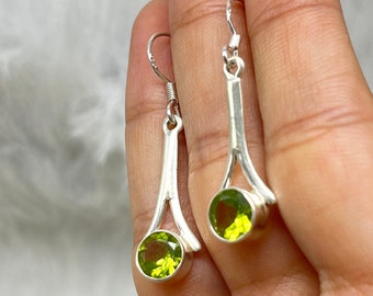 Puede incluir: Un par de pendientes colgantes de plata con detalles de piedras preciosas verdes. Los pendientes presentan un diseño simple y elegante con una sola piedra preciosa redonda en la parte inferior de cada pendiente.