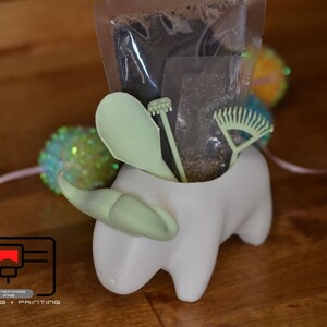 May include: A white ceramic planter shaped like a bull with a green horn. The planter contains a small bag of soil, a green trowel, a green rake, and a green gardening tool.