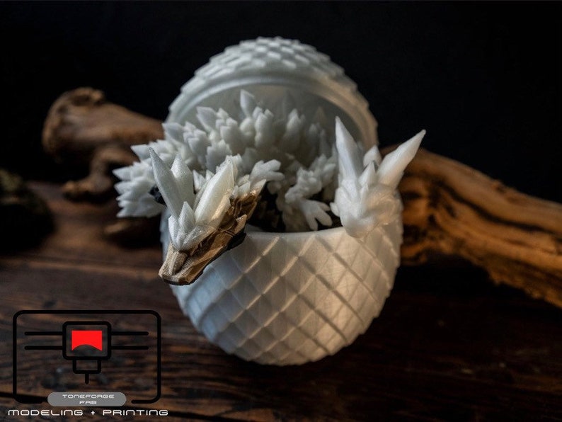 May include: A white 3D printed dragon sculpture emerging from a white egg-shaped container. The dragon has a spiky, crystalline texture and is partially visible from the open top of the egg.