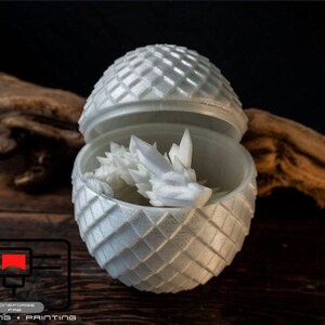 May include: A white 3D printed dragon egg with a dragon inside. The egg is a sphere with a textured surface and the dragon is curled up inside. The egg is open and the dragon is visible.