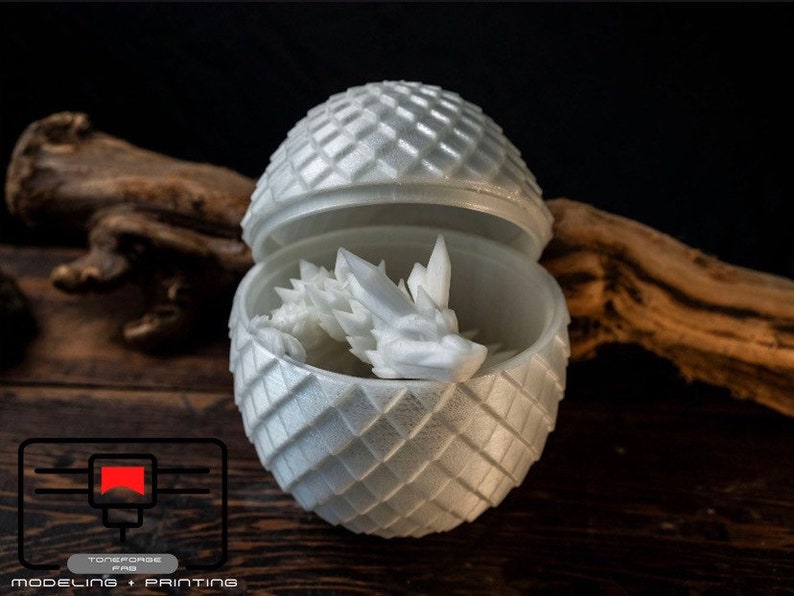 May include: A white 3D printed dragon egg with a dragon inside. The egg is a sphere with a textured surface and the dragon is curled up inside. The egg is open and the dragon is visible.