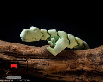 Puede incluir: Un modelo de dinosaurio articulado impreso en 3D de color verde, posado sobre una rama marrón. El dinosaurio tiene una cola larga y una cabeza pequeña con una sonrisa de dientes.