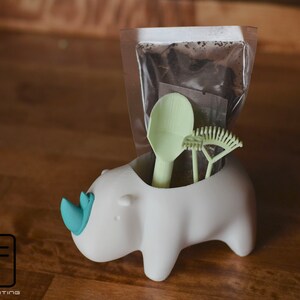 May include: A white rhinoceros-shaped planter with a light blue horn, holding a clear plastic bag with brown soil, a light green spoon, and a light green rake.