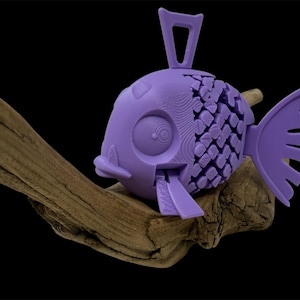 Puede incluir: Una figura de pez morado impresa en 3D con un cuerpo segmentado y articulado, ojos circulares grandes y una prominente aleta dorsal en forma de bucle. El pez presenta un patrón de escamas texturizado en el centro y descansa sobre madera flotante.