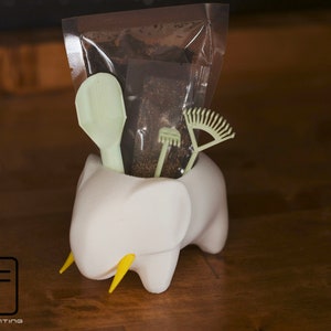 May include: A white elephant-shaped planter with yellow tusks, filled with a small bag of brown soil, a green scoop, a small green rake, and a green fan-shaped rake.