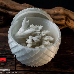 May include: A white 3D printed dragon egg with a lid open, revealing a white crystal-like object inside. The egg is sitting on a wooden surface.