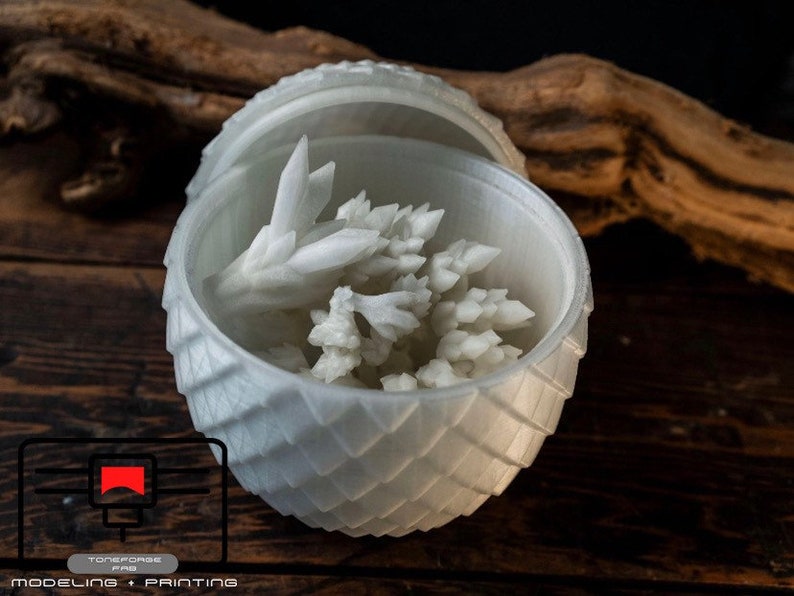May include: A white 3D printed dragon egg with a lid open, revealing a white crystal-like object inside. The egg is sitting on a wooden surface.