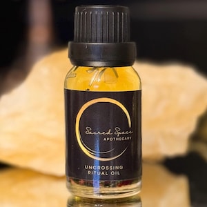 Könnte beinhalten: Eine kleine Glasflasche "Uncrossing Ritual Oil" von Sacred Space Apothecary. Die Flasche hat ein schwarzes Etikett mit goldfarbenen Buchstaben und einem goldenen Halbmond-Design. Das Öl im Inneren hat eine goldgelbe Farbe. Die Flasche spiegelt sich auf einer glänzenden schwarzen Oberfläche.