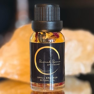 May include: A small glass bottle with a black cap and a dark label featuring gold text and a circular design. The label reads "Sacred Space Apothecary" and "Spell Breaker Ritual Oil." The bottle contains a clear liquid with visible botanicals.
