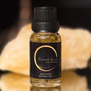 Può includere: Una piccola bottiglia di vetro di "Success Ritual Oil" con un'etichetta nera e scritte dorate. L'etichetta presenta un design circolare dorato e le parole "Sacred Space Apothecary". La bottiglia è riempita di un liquido dorato.