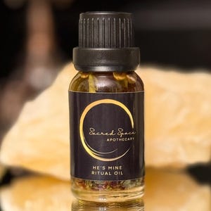 May include: A small glass bottle of "He's Mine Ritual Oil" with a black cap and a dark label featuring gold lettering and a circular design. The bottle is filled with a golden liquid and small herbs.