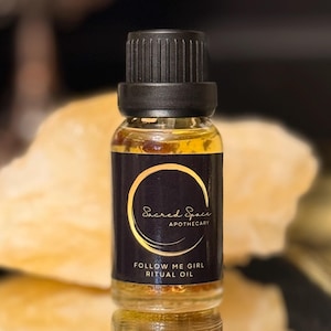 Può includere: Una piccola bottiglia di vetro di olio rituale con tappo nero. L'etichetta nera ha scritte dorate che recitano "Sacred Space Apothecary" e "Follow Me Girl Ritual Oil". L'olio è color ambra.