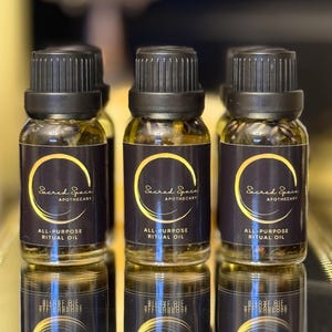 Può includere: Tre piccole bottiglie di vetro di Olio Rituale Multiuso con tappi neri ed etichette scure. Le etichette presentano testo dorato e un design circolare dorato con le parole "Sacred Space Apothecary". Le bottiglie si riflettono su una superficie lucida.