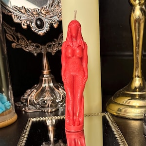 May include: A red candle sculpted as a female form. The candle is a vibrant red and has a detailed design. It stands on a mirrored surface, with other decorative items visible in the background.