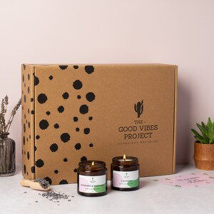 May include: Two brown glass candle jars with lids. The candle on the left is labeled "Mandarin &  Blood Orange" and the candle on the right is labeled "Lavender". The candles are in front of a brown cardboard box with black polka dots and the text "The Good Vibes Project - Conscious Wellbeing".