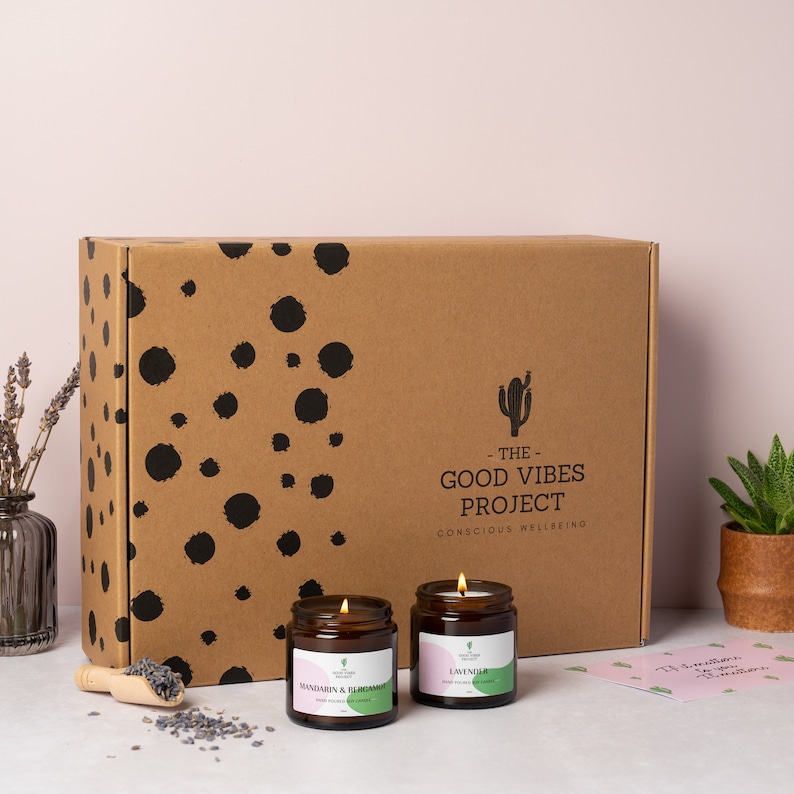May include: Two brown glass candle jars with lids. The candle on the left is labeled "Mandarin &  Blood Orange" and the candle on the right is labeled "Lavender". The candles are in front of a brown cardboard box with black polka dots and the text "The Good Vibes Project - Conscious Wellbeing".