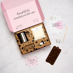 May include: An open pink box with a brown and black patterned interior. The box contains a soy wax melt making kit with a metal pitcher, a bag of soy wax flakes, a small bottle of essential oil, a wooden stirring stick, and a silicone mould. The box also includes a small card with the text "I hope you knew the world is better because you are in it."  The box is labelled "thoughtfully curated just for you".