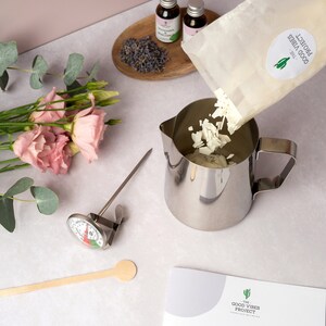 May include: A white bag of soy wax flakes is being poured into a stainless steel pitcher. A thermometer, wooden spoon, and essential oil bottles are also in the image. The bag has the text "The Good Vibes Project" printed on it.