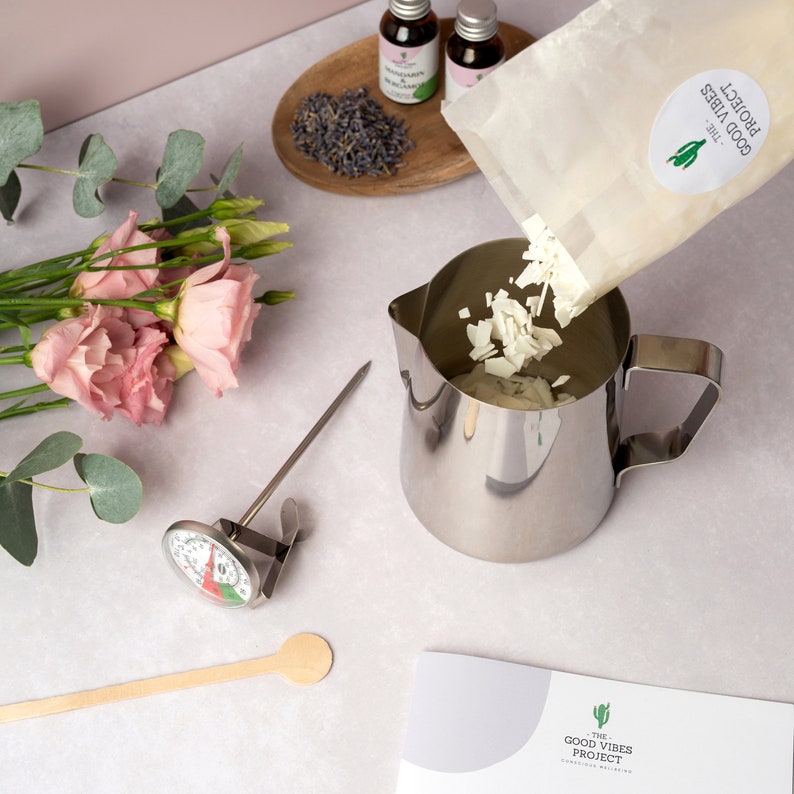 May include: A white bag of soy wax flakes is being poured into a stainless steel pitcher. A thermometer, wooden spoon, and essential oil bottles are also in the image. The bag has the text "The Good Vibes Project" printed on it.
