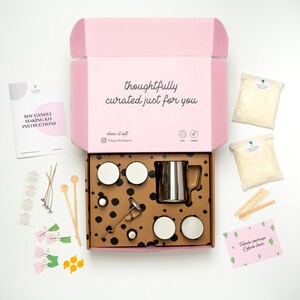May include: A pink box with the text "thoughtfully curated just for you" on the lid. The box contains a soy candle making kit with all the necessary supplies, including soy wax flakes, wicks, jars, a thermometer, and a pouring pitcher. The kit also includes a small card with the text "Inhale courage Exhale fear".