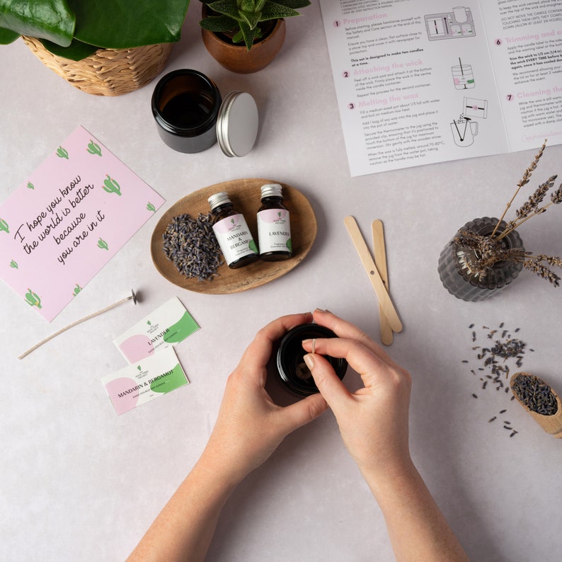 May include: A candle making kit with two small glass bottles of essential oils, lavender and mandarin & bergamot, a wooden tray with dried lavender, two wooden stirring sticks, a black candle jar, a wick, and two small cards with the words "Lavender" and "Mandarin & Bergamot" printed on them.