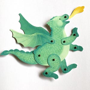 May include: A green paper dragon cutout with articulated limbs and wings attached by small metal brads. The dragon features a textured scale pattern in shades of light and dark green, with a yellow flame shape emerging from its open mouth.