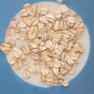 May include: A close-up shot of oatmeal flakes in a creamy liquid, set against a blue background. The oatmeal is a light tan color, and the liquid is white. The image is well-lit and shows the texture of the oatmeal.