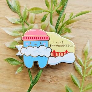 May include: A blue and pink enamel pin featuring a cartoon character wearing a red hat and a cloud. The character is standing in front of a yellow and red depiction of the Golden Gate Bridge. The text "I love San Francisco" is written on the pin.