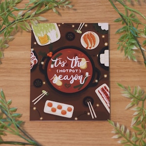 May include: A festive greeting card featuring a hot pot illustration. The card depicts a red hot pot with various ingredients, including vegetables, meat, and dumplings. The text on the card reads "'Tis the (hotpot) season".