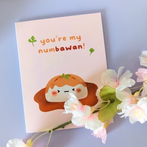 Puede incluir: Una tarjeta rosa con el texto "you're my numbawan!" y una ilustración de dibujos animados de un postre marrón y blanco con una ramita verde en la parte superior.
