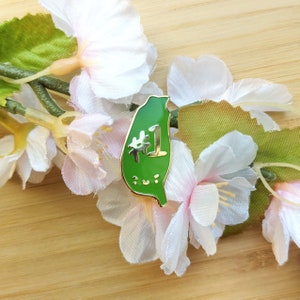 Puede incluir: Un pin de esmalte verde con forma de Taiwán, con borde dorado y el número "1" y un diseño floral. El pin está colocado sobre un fondo de flores blancas y rosas y hojas verdes.