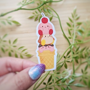 Könnte beinhalten: Ein Aufkleber mit einem Stapel von drei Cartoon-Charakteren, die Eiscremekugeln ähneln, gekrönt von einer Kirsche, auf einer Waffel. Der Aufkleber hat ein hellrosa, braunes und gelbes Farbschema, mit dem Text "blueberry art" auf der Waffel.