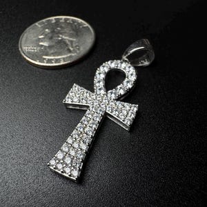 May include: A silver-colored ankh pendant encrusted with numerous small, clear gemstones. The pendant is displayed next to a U.S. quarter for size comparison. The ankh is a symbol of life and immortality.