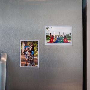May include: Two framed photos on a stainless steel refrigerator. The top photo shows a group of children wearing superhero capes and standing with their arms raised. The bottom photo shows a group of children in a park setting.