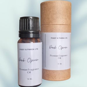 Op de afbeelding: Een kleine glazen fles "Dark Opium" premium geurolie met een zwarte dop, naast een cilindrische kartonnen container. Beide items hebben witte labels met zwarte tekst, waaronder het merk "Piggy & Panda LTD" en het volume van 10 ml.