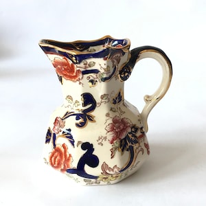 Op de afbeelding: Een witte keramische kan met een blauw en gouden bloemenpatroon. De kan heeft een gegolfde rand en een handvat.