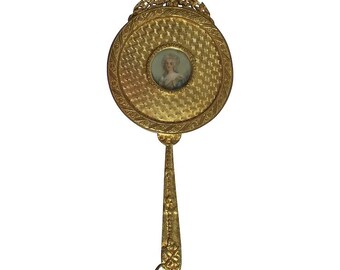 May include: A gold-toned hand mirror with a round, ornate frame and a small portrait of a woman in the centre. The handle is decorated with floral details.