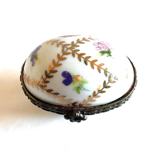 May include: A white porcelain trinket box with a domed lid featuring a gold leaf lattice pattern and small hand-painted floral motifs in pink, purple, and green. The base is encased in a dark, textured metal frame with a visible hinge mechanism.