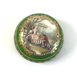 Puede incluir: Una pequeña caja de porcelana redonda de color verde con un borde dorado y una escena pintada de una pareja en un jardín.