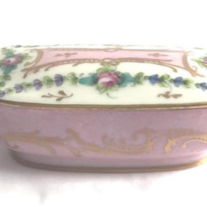 May include: A small, oval-shaped porcelain trinket box with a pink base and a white lid. The lid is decorated with a floral design in shades of pink, blue, and green, and a gold trim.