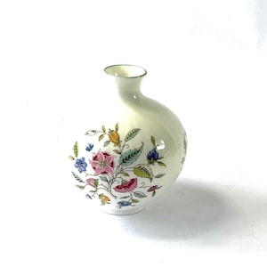 May include: A small, off-white ceramic vase with a bulbous body and a slender neck. The vase is adorned with a colorful floral pattern featuring pink, blue, and yellow flowers with green leaves. The rim is trimmed with green.