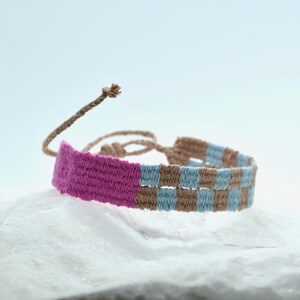 Op de afbeelding: Een geweven vriendschapsarmband met een effen magenta gedeelte gevolgd door een herhalend ruitpatroon in lichtblauw en beige. De armband is voorzien van dunne, verstelbare gevlochten koordjes aan de achterkant om het item rond de pols te bevestigen.