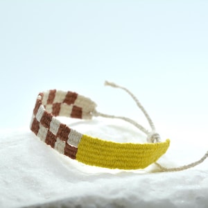 Op de afbeelding: Een geweven armband met een ruitpatroon in bruin en gebroken wit aan de ene kant en een effen felgeel gedeelte aan de andere kant. De armband is voorzien van dunne, lichtgekleurde touwtjes aan beide uiteinden om het sieraad vast te maken.