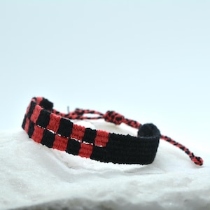 Op de afbeelding: Een geweven armband met een herhalend dambordpatroon van zwarte en rode draad. Het ontwerp gaat over in een effen zwart gedeelte, met dunne gevlochten koordjes aan de uiteinden voor een verstelbare sluiting rondom de pols.