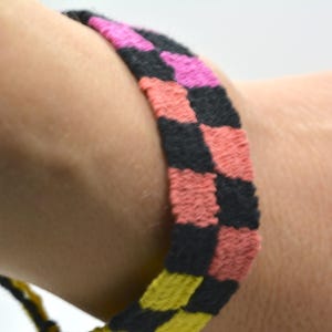 Op de afbeelding: Een close-up van een geweven armband met een dambordpatroon. De armband heeft zwarte, roze en koraalkleurige vierkanten. De armband wordt om een pols gedragen. De armband is een handgemaakt accessoire.