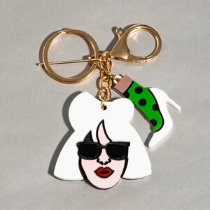 Può includere: Un portachiavi color oro con un volto bianco in stile cartone animato con occhiali da sole neri e rossetto rosso. È attaccato un ciondolo a forma di calzino verde a pois neri e un tacco alto bianco.