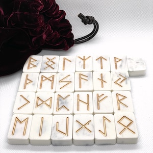 Puede incluir: Conjunto de 24 piedras de runas de mármol blanco con runas doradas. Las runas están dispuestas en cuatro filas de seis piedras cada una. Las piedras están sobre una superficie blanca.